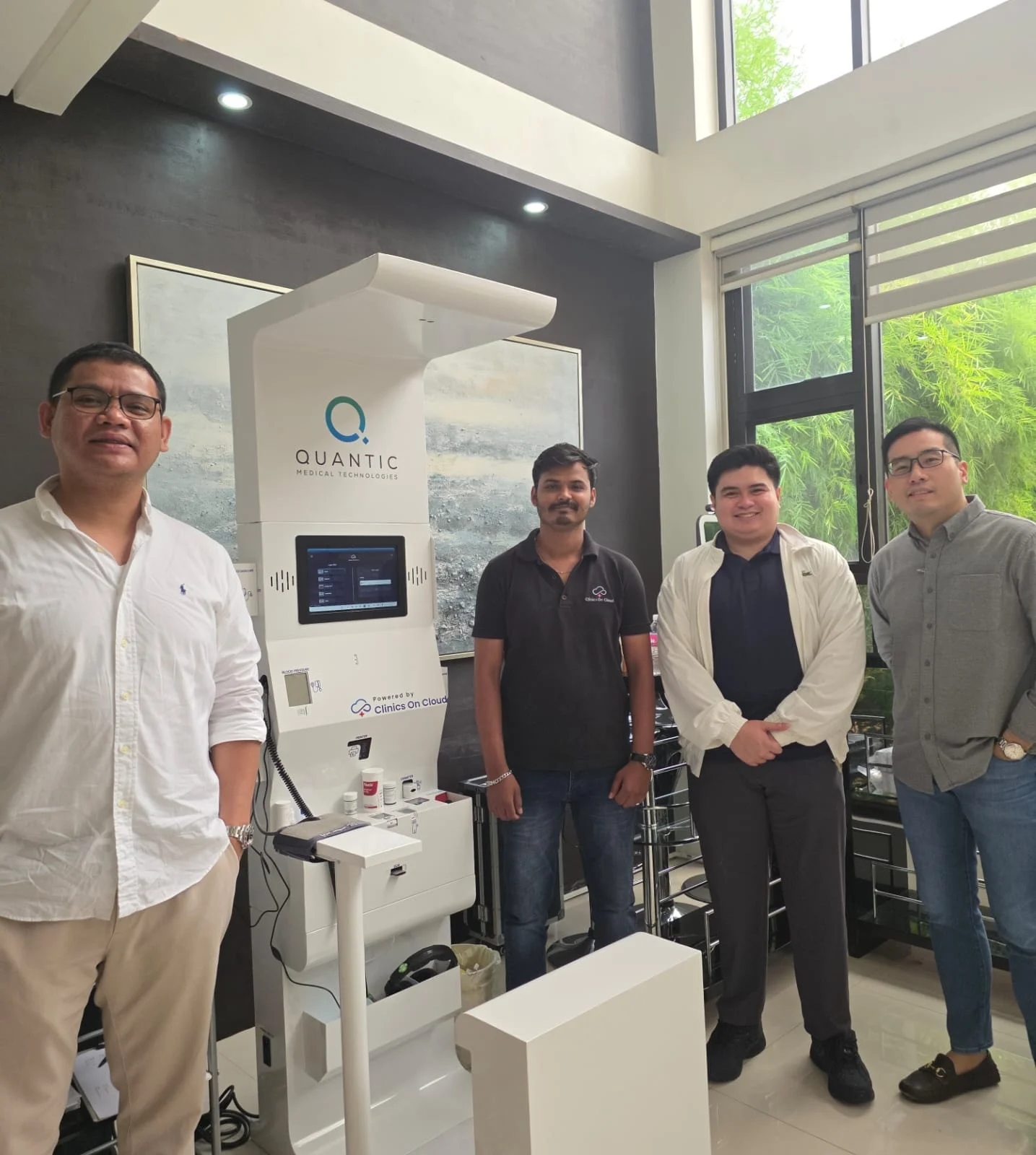 Four men stand beside a Quantic health kiosk with touchscreen in a bright indoor space with tall windows.
